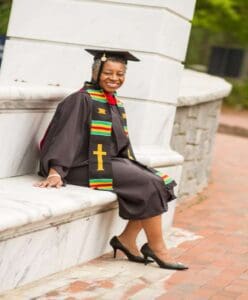 GRADUATE SUNDAY 2021 Carol Tucker Burden A woman in graduation attire sitting on the side of a wall.