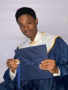 A young man in graduation attire holding his cap and gown.