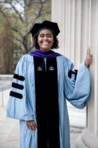 A woman in graduation gown and cap standing next to a pillar.