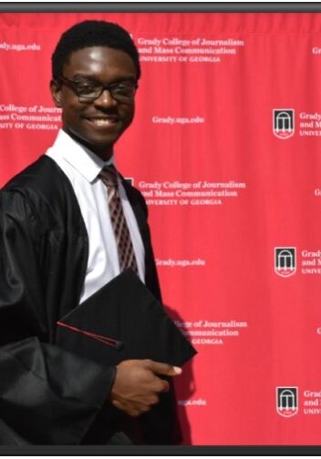 A man in graduation attire standing next to a red wall.